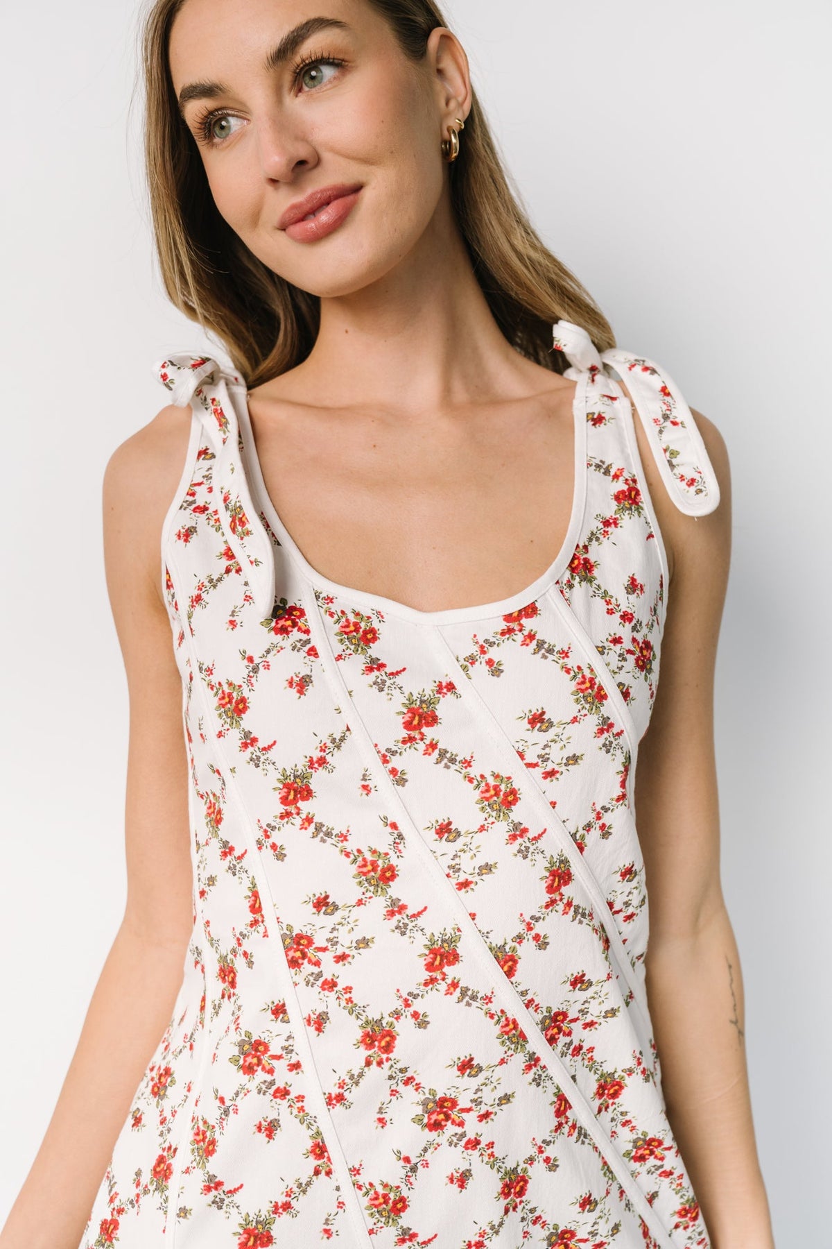 Three-quarter view showing fitted waist, flowing skirt, and the right-side slit on an off-white dress with red floral pattern and bow-tie straps.