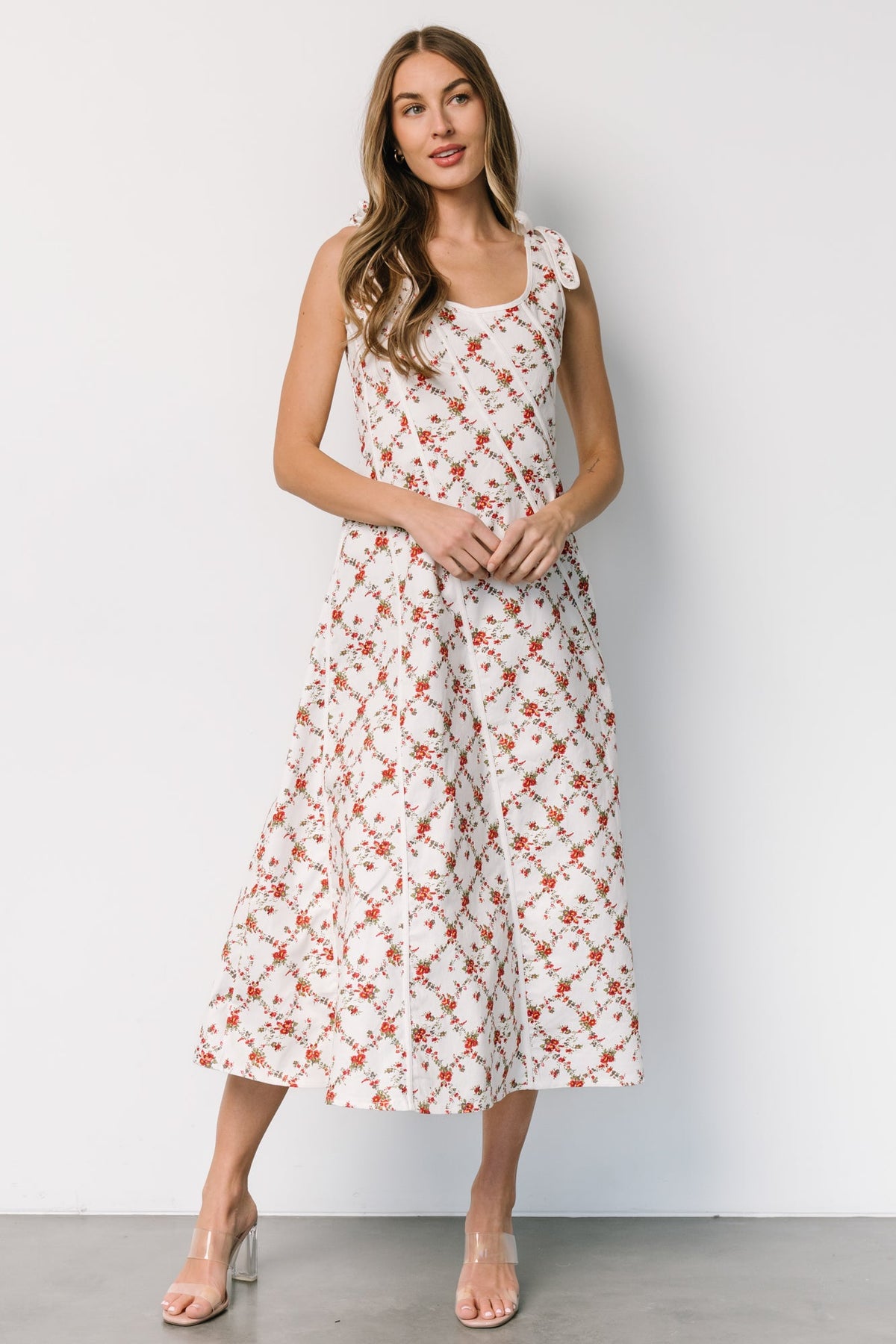 Front view of off-white midi dress with red and green florals, scoop neckline, bow tank straps, diagonal piping, and a right-side slit for movement.