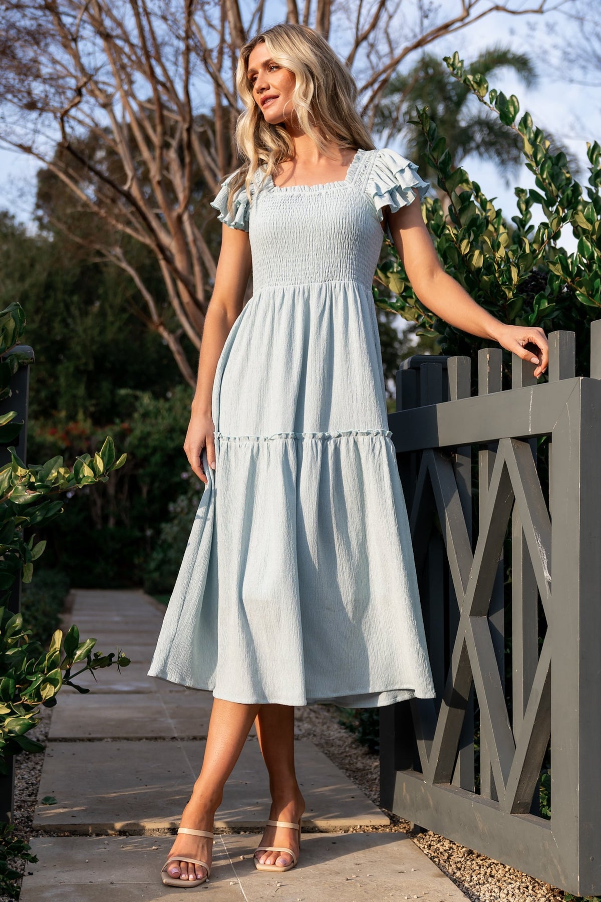 Back view highlighting the smocked back bodice of the light blue midi dress and the smooth single-tier skirt with hidden side pockets.
