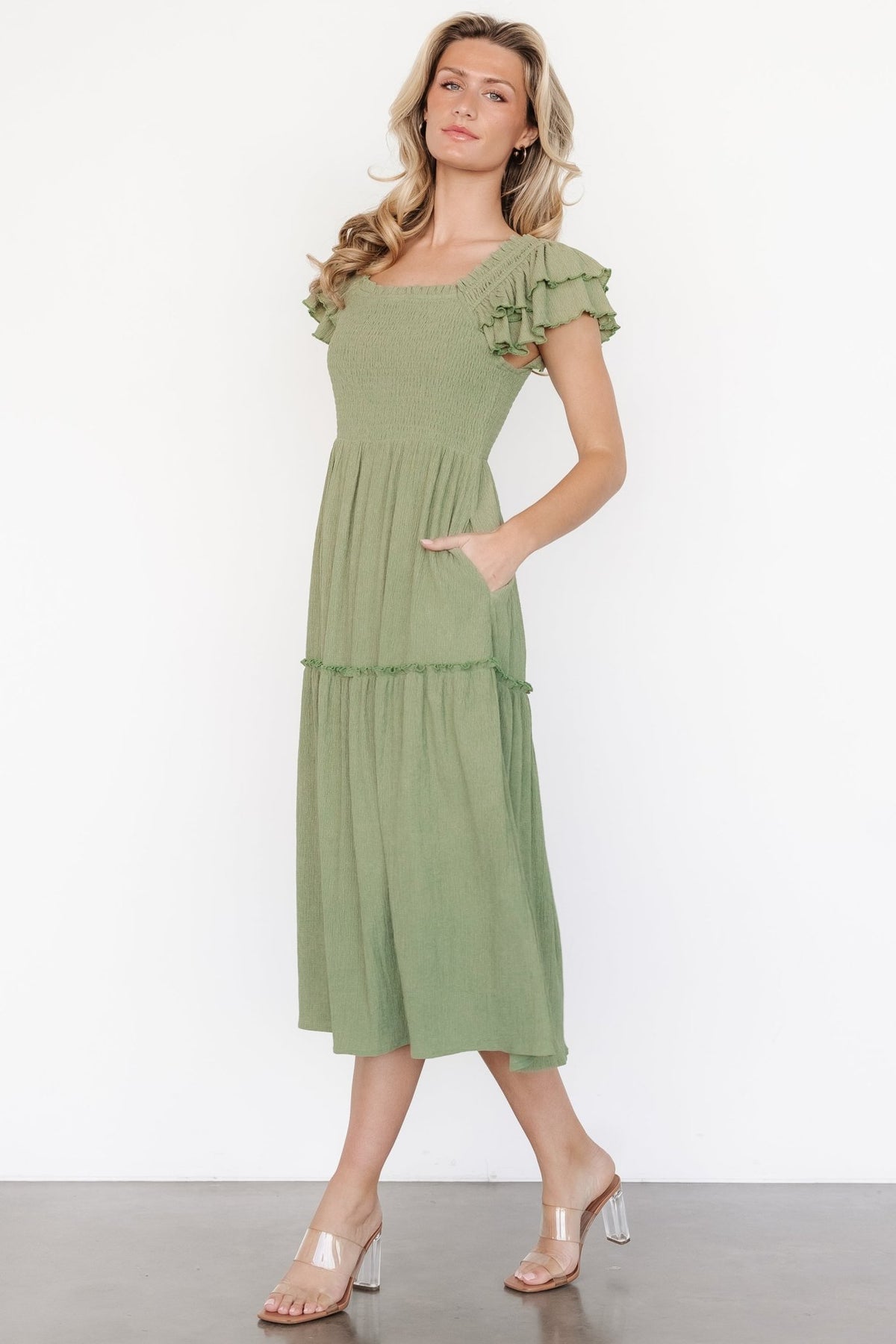 Front view of model wearing the Jacie Smocked Midi Dress in sage green with square neckline, smocked bodice, ruffle cap sleeves, and pockets, studio shot.
