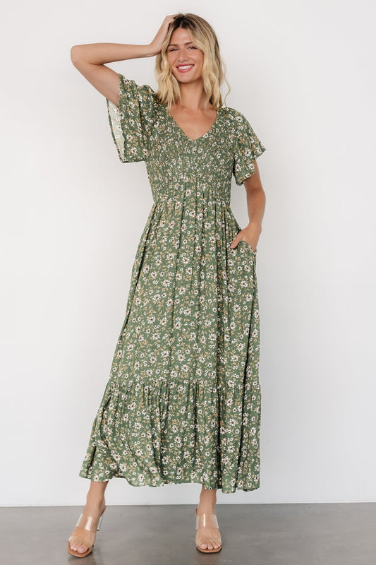 Full-length view of model wearing green floral smocked midi dress with flutter sleeves, smocked bust, empire waist, and hidden side pockets.