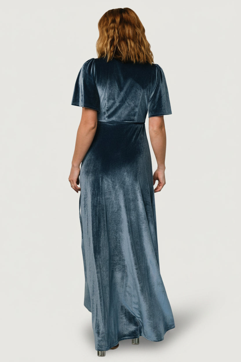 Model wearing Katelyn velvet wrap dress in blue walking to show movement and how the tulip skirt reveals subtle leg.