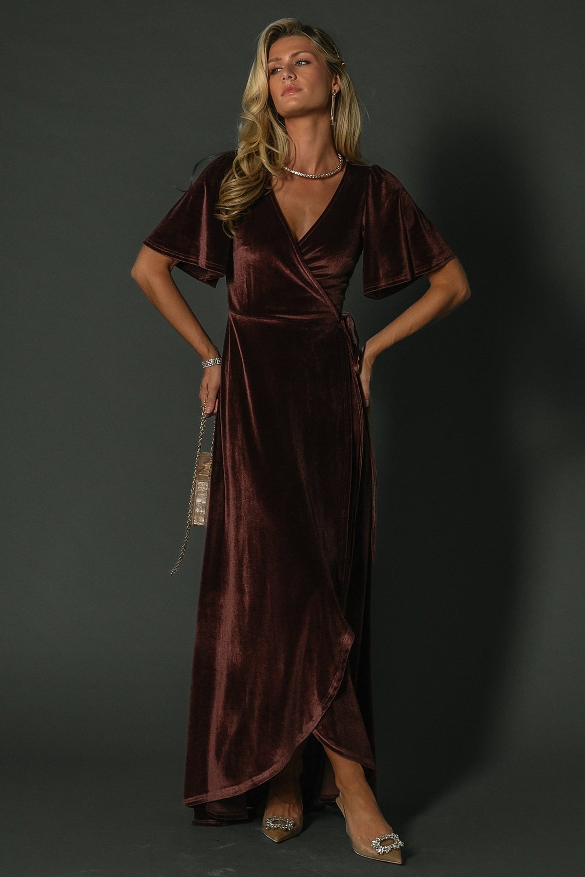 Full-length front view of Katelyn Velvet Maxi Wrap Dress in clove on model, showing tulip skirt, medium flutter sleeves, and V-neck wrap silhouette.