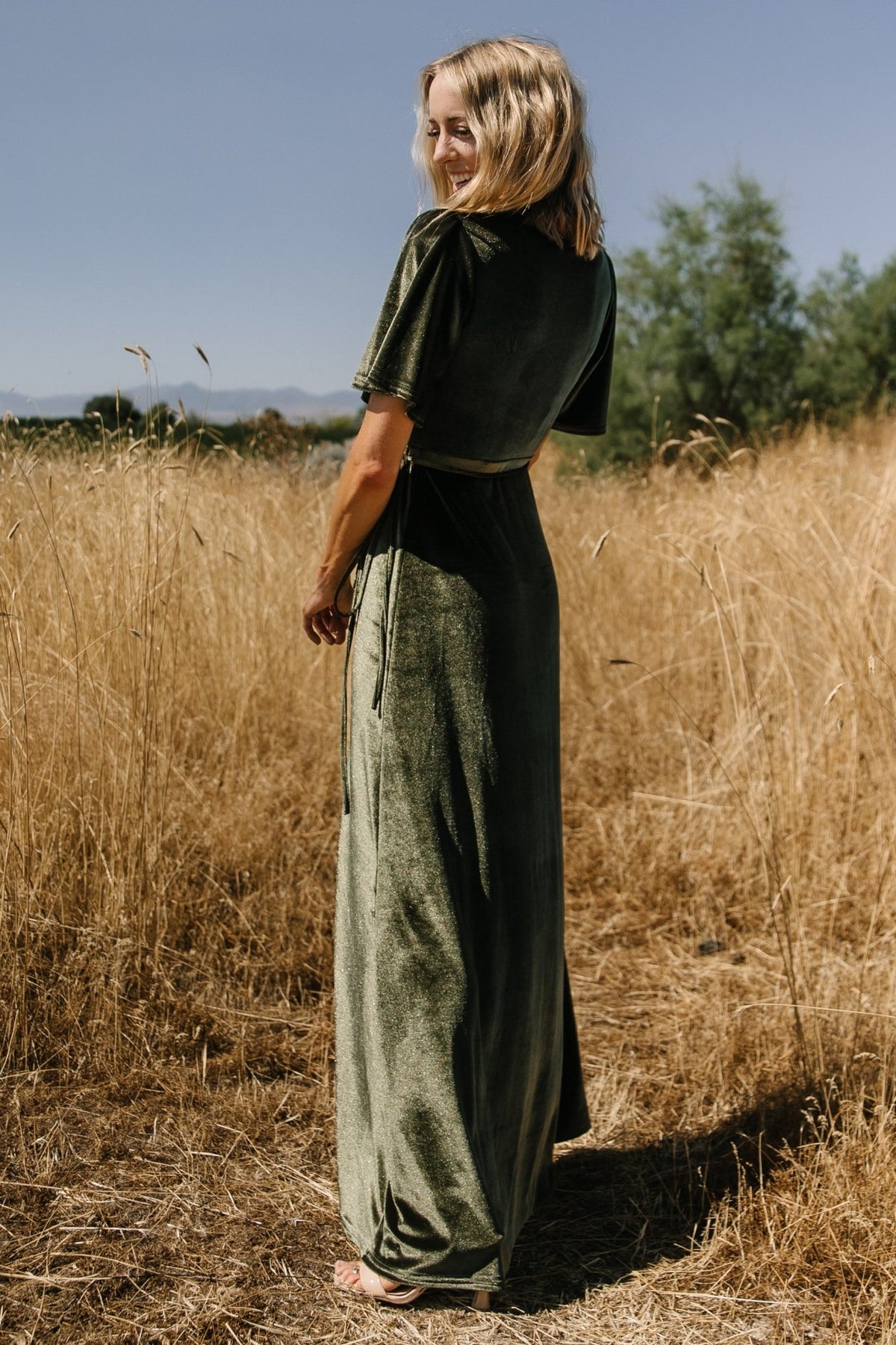 Full-length view of the dark olive Katelyn Velvet Maxi Wrap Dress on model, illustrating the tulip skirt slit and elegant silhouette.