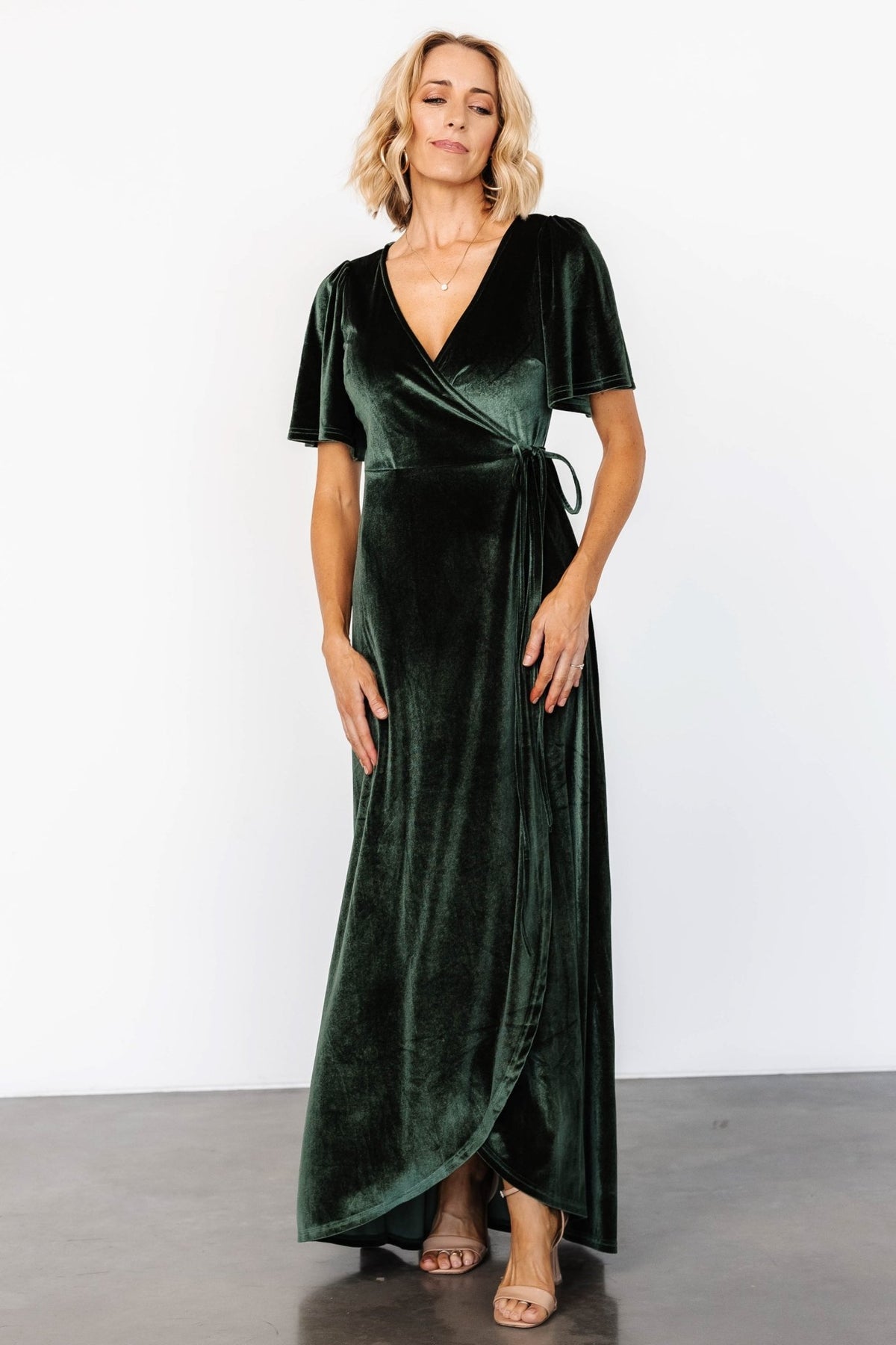 Side angle of deep green velvet wrap dress highlighting the waist tie loop, tulip skirt overlap, and soft velvet drape.