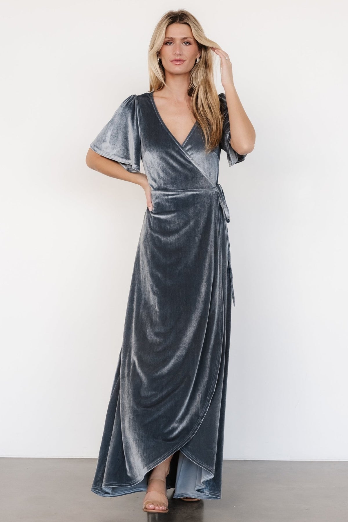 Front view of dusty blue velvet wrap maxi dress on model showing V-neck, flutter sleeves, and tulip skirt with natural drape and movement, fully lined.