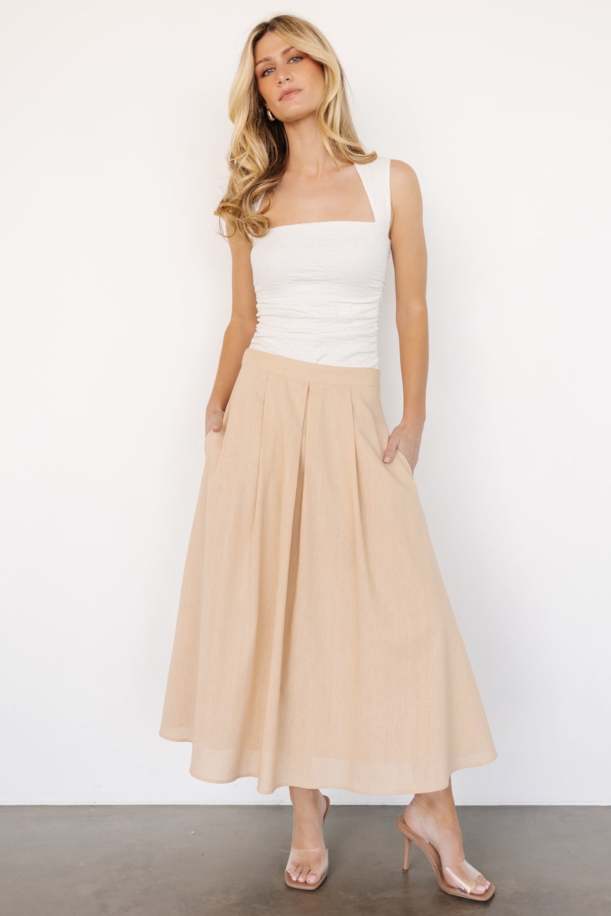 Model wearing natural pleated midi skirt styled with a tucked top, showing mid-calf length, hidden side pockets, and pleat movement.