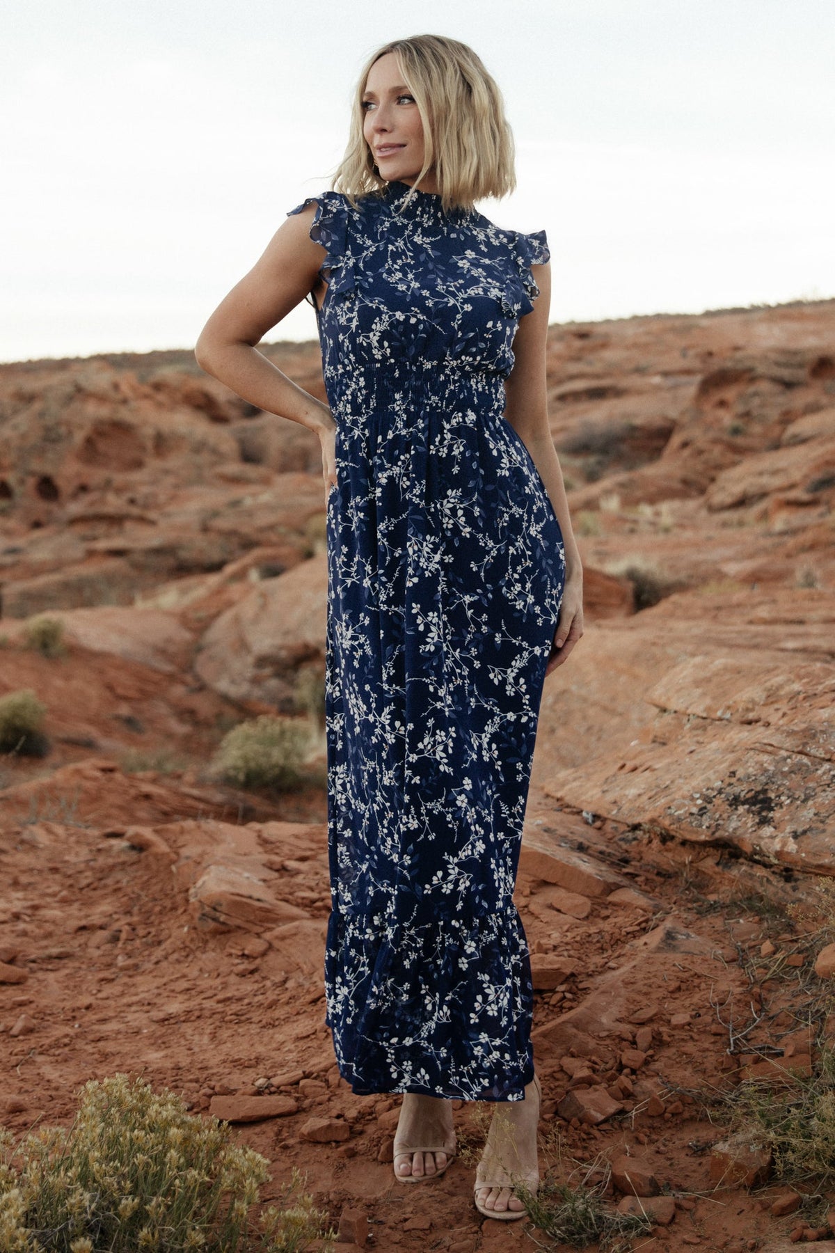 Front full-length view of blue floral chiffon maxi dress with smocked high neckline, short flutter sleeves, ruffle hem, and lined skirt on a model.