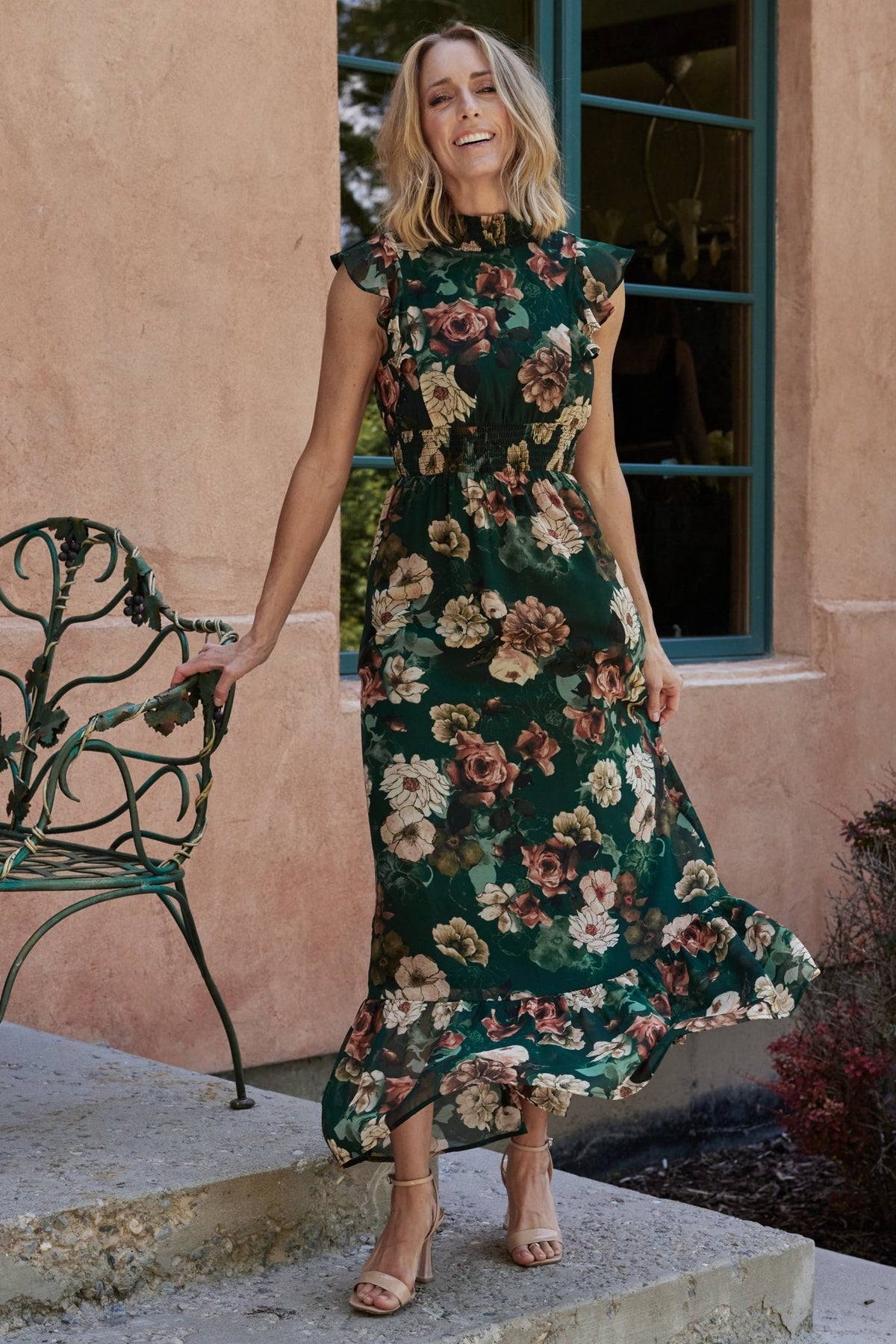 Dark green floral chiffon maxi dress on model; smocked high neckline, short flutter sleeves, ruffle hem and full-length flow visible.