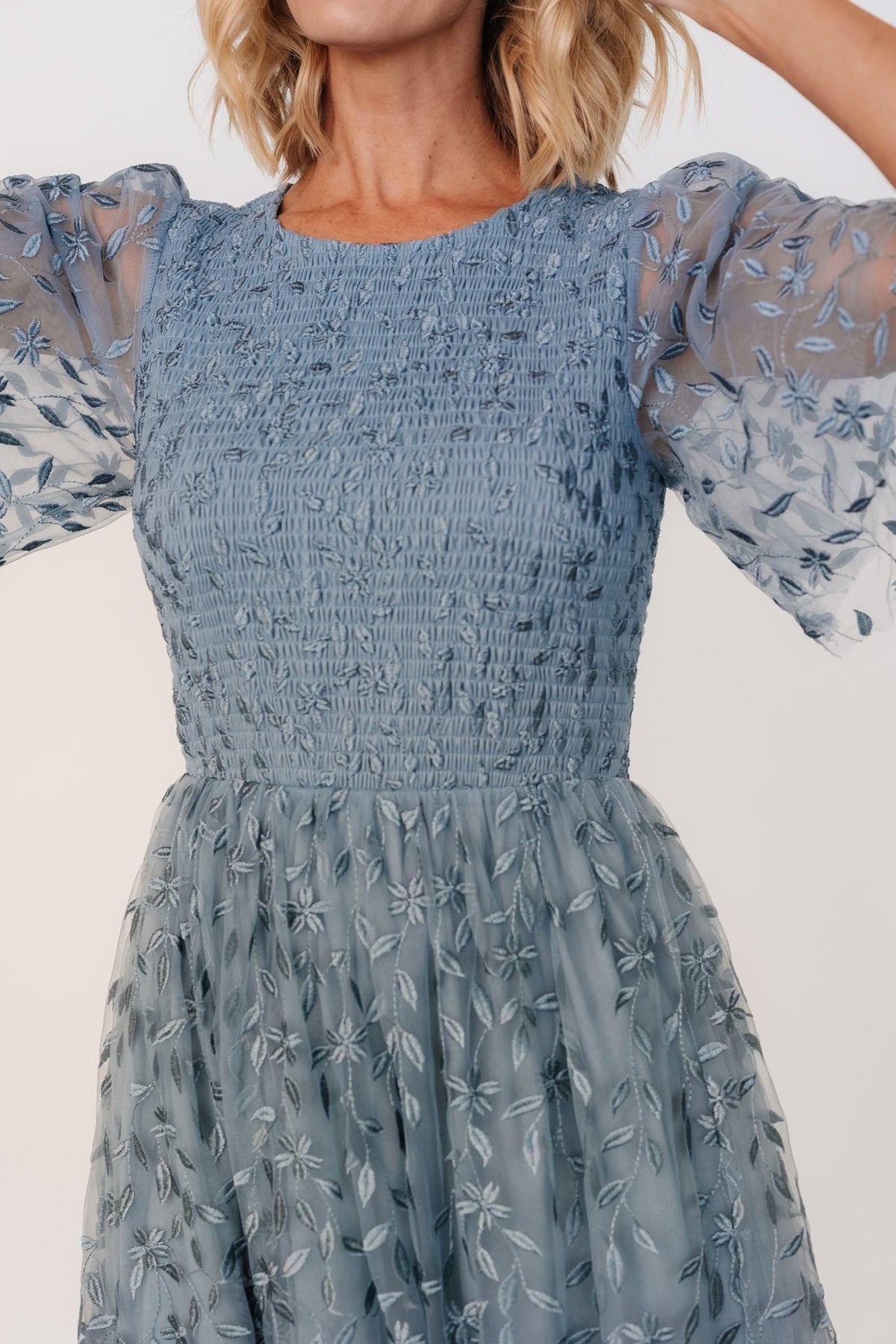 Model wearing dusty blue Kendra dress showing flutter-style tulle sleeves, round neckline and smocked bodice for a fitted, comfortable look.