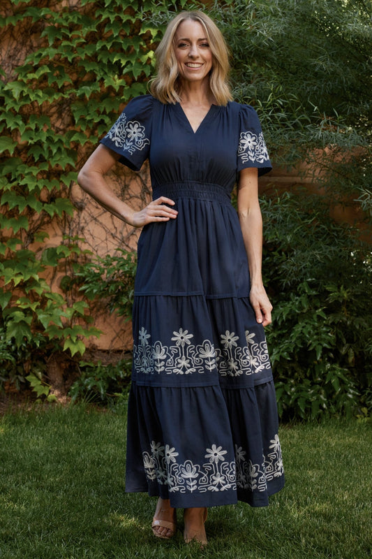 Full-length front view of blue embroidered maxi dress with smocked waist, short sleeves, tiered skirt and side pockets, on model.