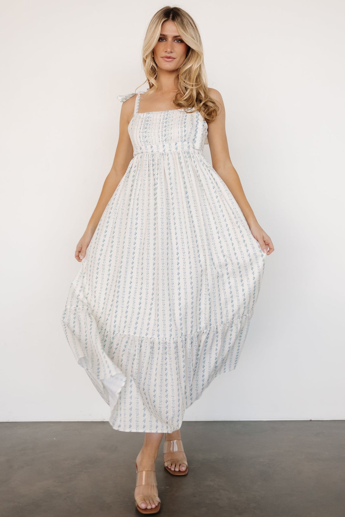 Model wearing Kennie Maxi Dress in cream multi floral showing relaxed fit, adjustable shoulder ties, left-side zipper, and flowing single-tiered skirt.