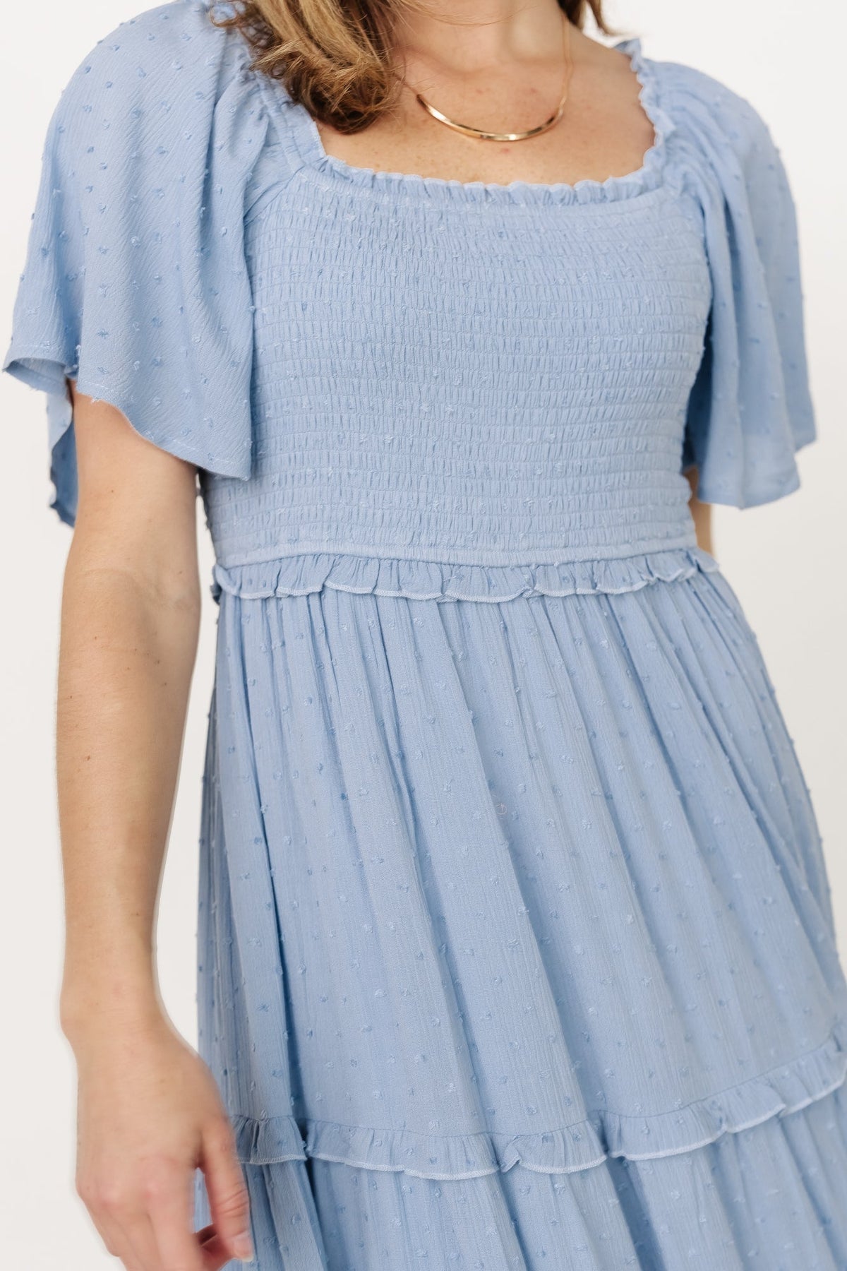 Back view of dusty blue Lenora dress showing smocked back detail, ruffled waist seam, and flowing tiered skirt to ankle length.