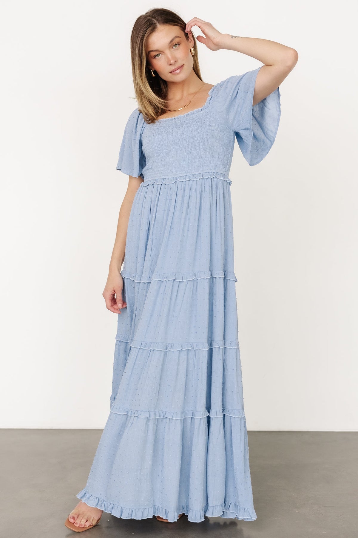 Model wearing dusty blue Lenora smocked dotted maxi dress showing elastic shoulders, short flutter sleeves, and defined ruffled waistline.