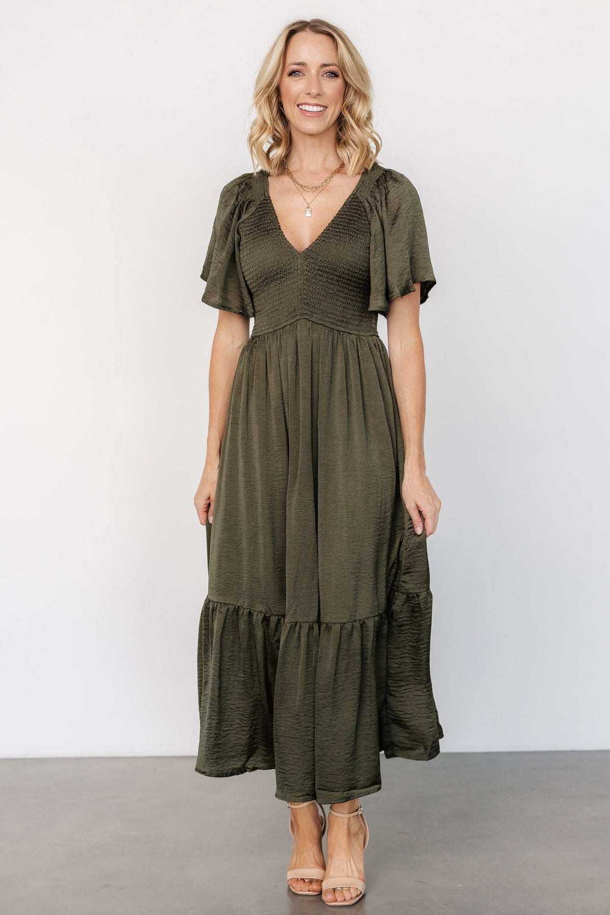 Three-quarter model view of Lovell smocked midi dress in deep olive highlighting satin-like sheen, fitted smocked top, and flowing skirt.