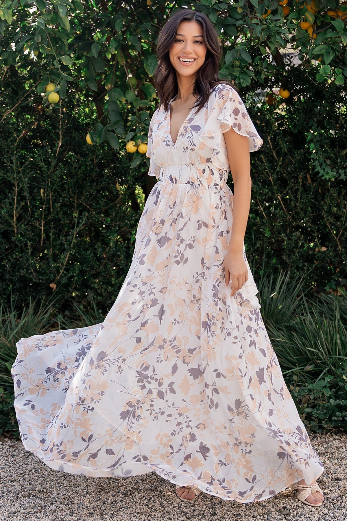 Off-white printed chiffon maxi dress with metallic floral shimmer, V-neckline, short flutter sleeves, and smocked waist; studio front view on model.