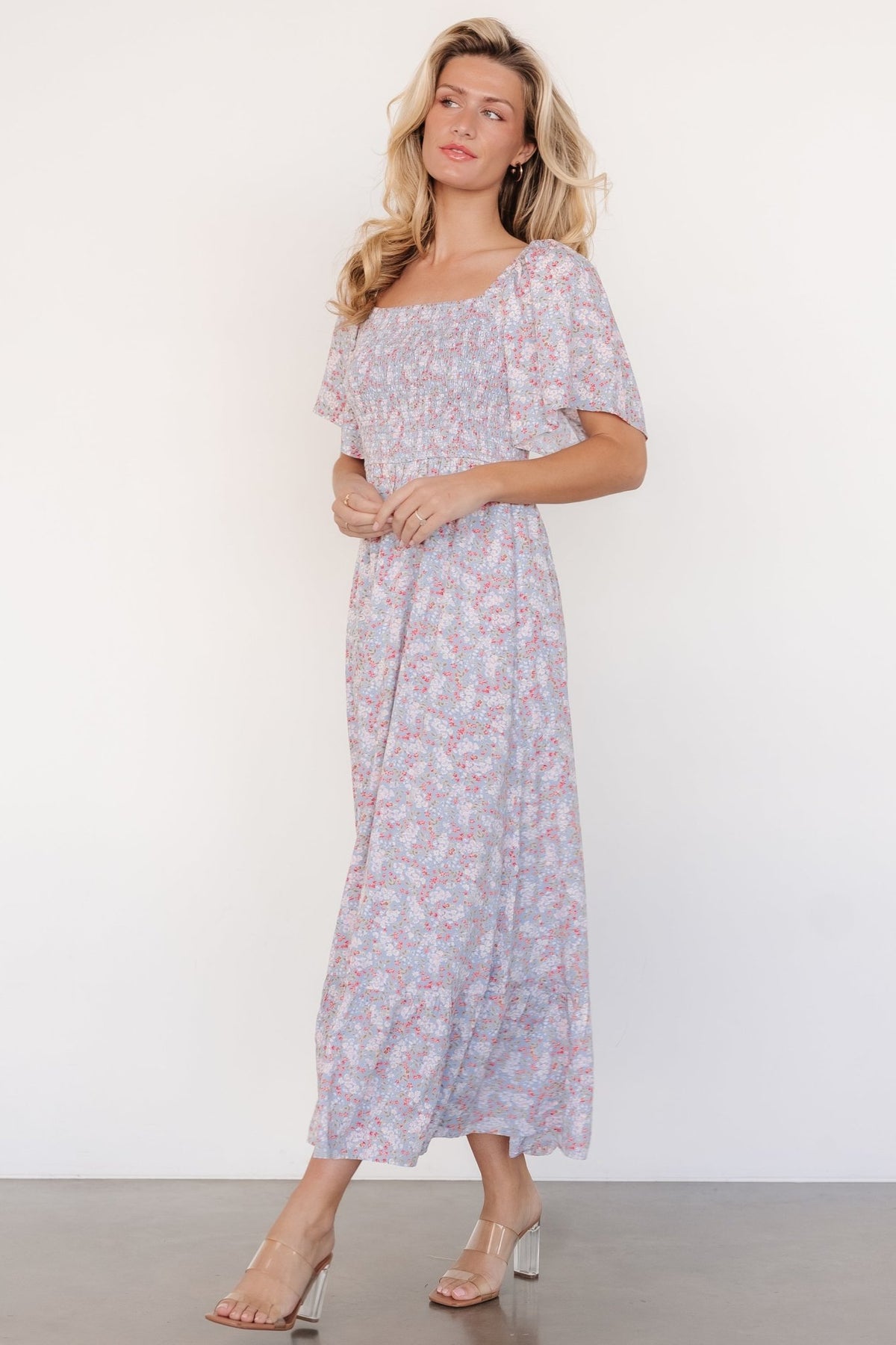 Model seated wearing Lyssa smocked midi dress in dusty blue floral, demonstrating comfortable movement, elastic waistline, and hidden side pocket placement.