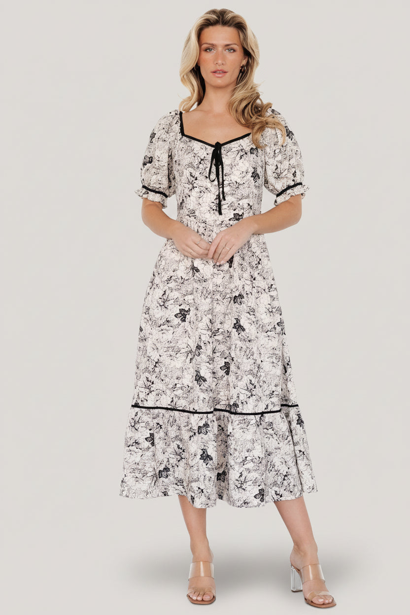 Front view of Mariana Midi Dress in cream and black floral on a model, showing sweetheart neckline, decorative bow, puff sleeves, and midi skirt.
