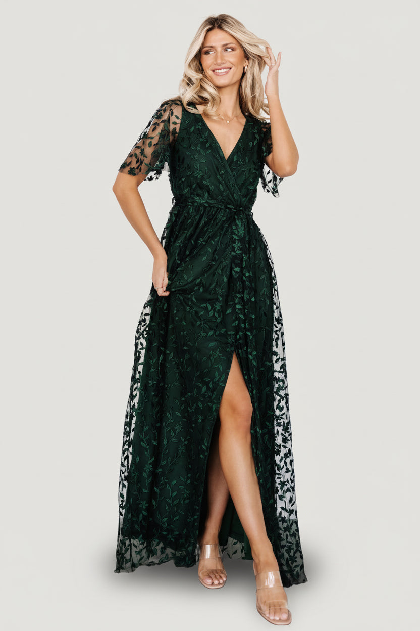 Full-length front view of dark green vine-embossed maxi dress with surplice neckline, elastic waistband, self-tie, and left-leg slit on a model.