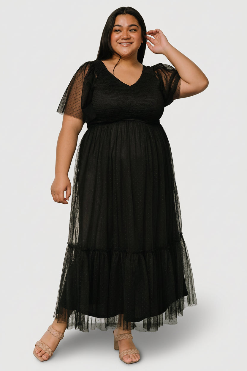 Model wearing black smocked tulle midi dress showing fitted smocked bodice, angled front waistline and knee-length tiered ruffle hem.