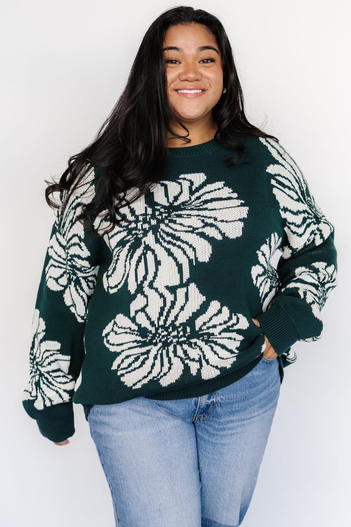Close-up of the Maude sweater showing medium-weight knit texture, wide ribbed cuff and off-white floral detailing on a deep green background.