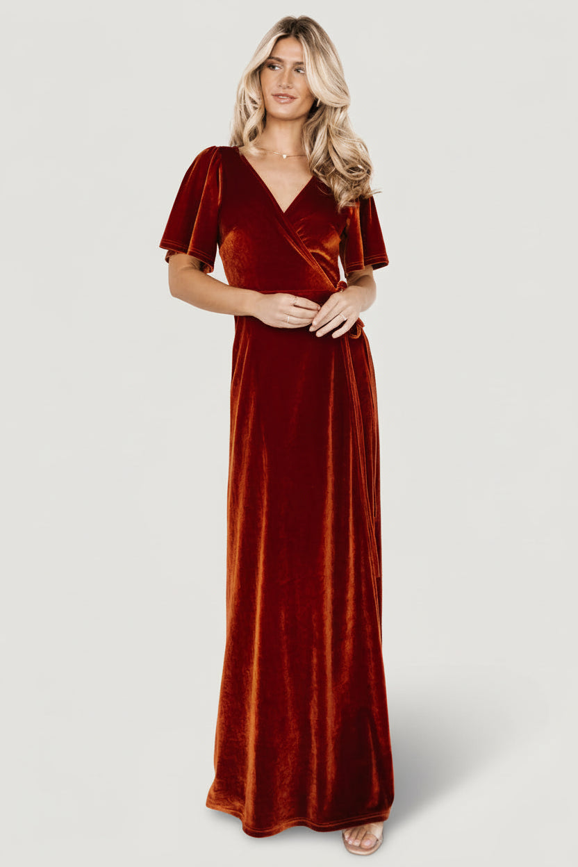 Front view of rust velvet wrap maxi dress on model; V-neck wrap bodice, self-tie waist, mid-length bell sleeves and tulip hem.
