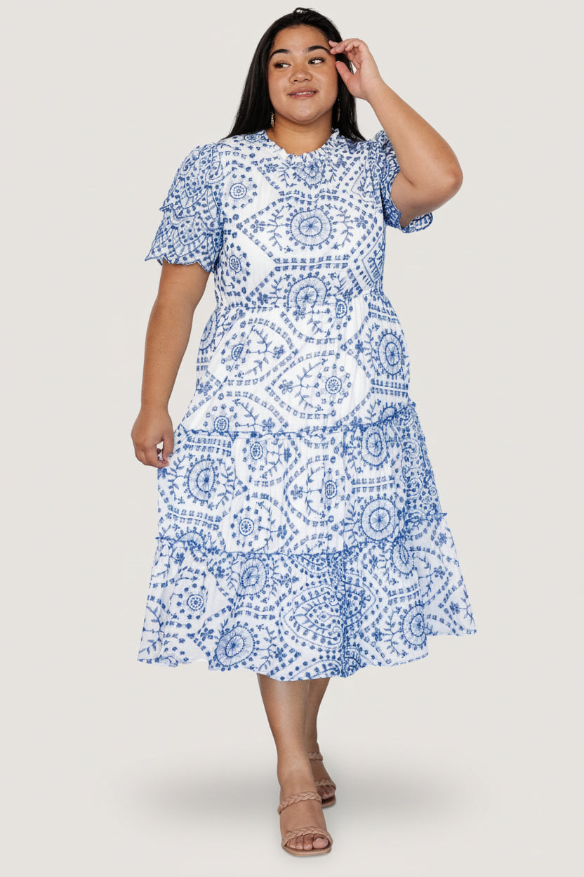 Back view showing keyhole and button closure, elastic waistband, and hidden side pocket on white midi dress with blue eyelet embroidery.