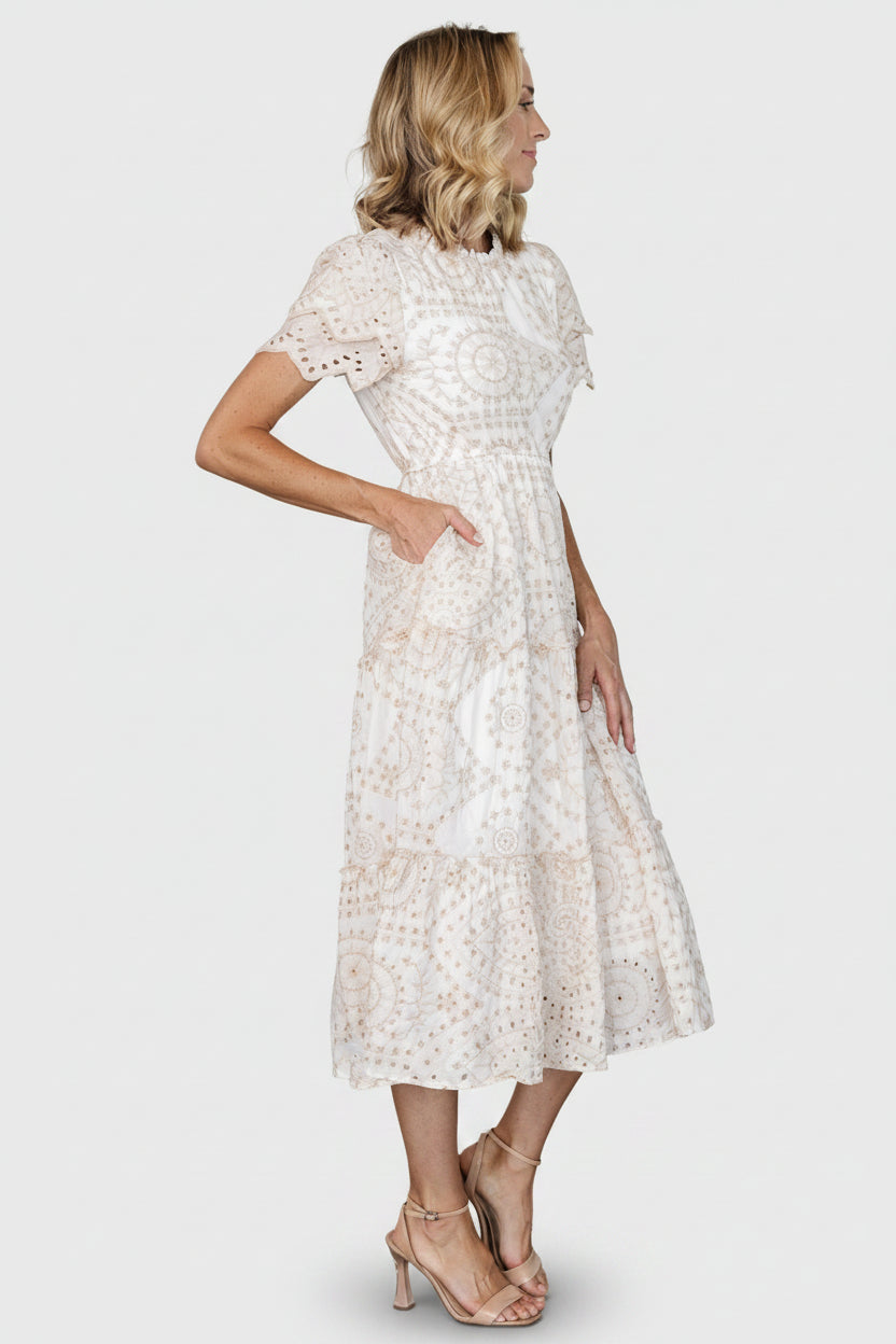 Close-up of natural beige eyelet embroidery on white cotton fabric, highlighting scalloped sleeve edges and ruffle detailing at the skirt tiers.