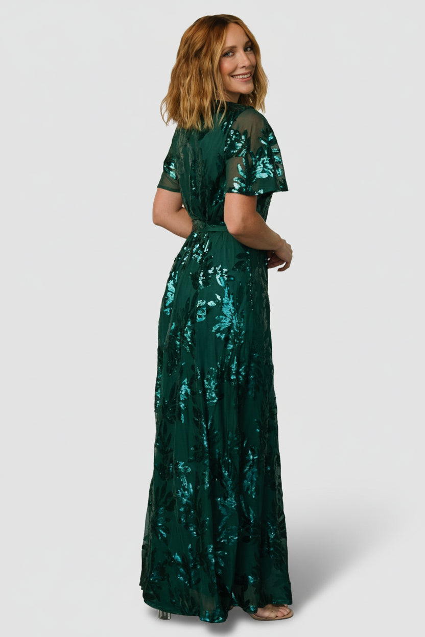 Side profile of Muse Sequin Maxi Dress in emerald green, illustrating fit through the waist, sash detail, and movement of the skirt.