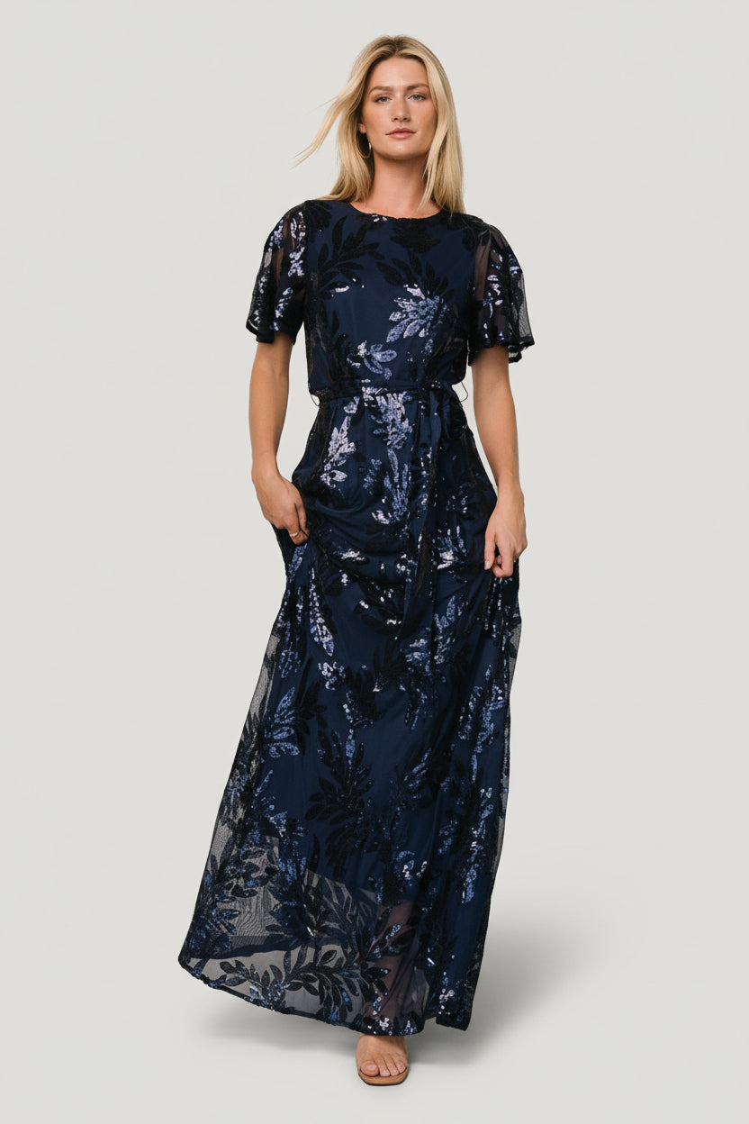 Front view of Muse Sequin Maxi Dress in navy with monochrome sequin leaf pattern, round neckline, waist sash, and full maxi silhouette.
