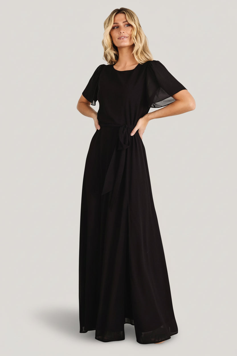 Front view of black chiffon Naomi maxi dress on model, showing rounded neckline, flutter sleeves, removable waist tie, and graceful drape.