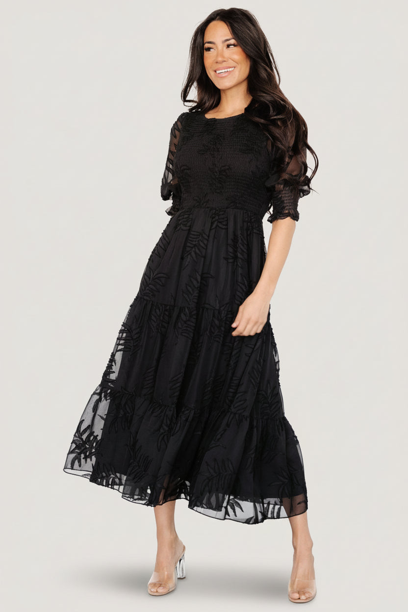 Full-length view of model wearing Nellie smocked midi dress in black chiffon with burnout botanical print, tiered skirt, and puff sleeves, standing straight.