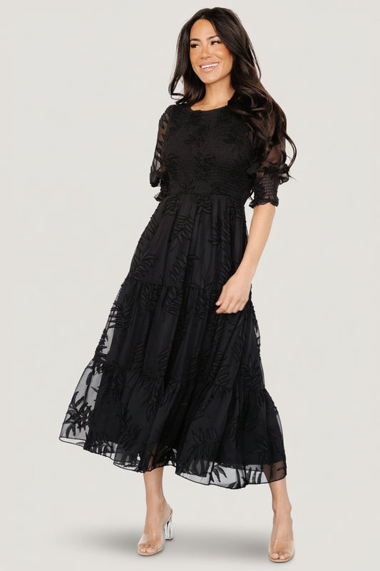 Full-length view of model wearing Nellie smocked midi dress in black chiffon with burnout botanical print, tiered skirt, and puff sleeves, standing straight.