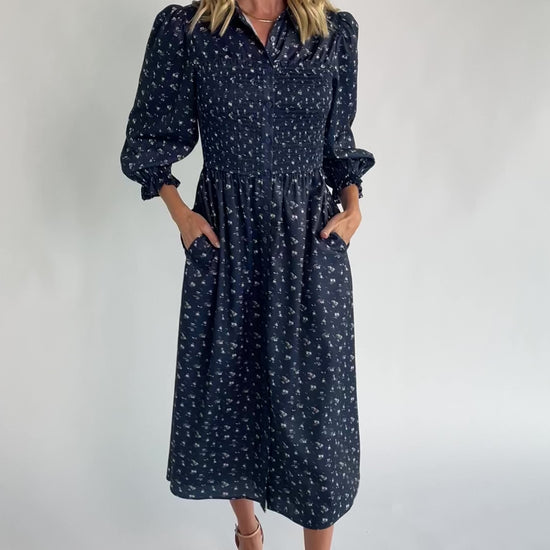 Navy floral button-down midi dress with a collared neckline, smocked bodice, three-quarter sleeves, elastic back waist, side pockets, and a flowing skirt.