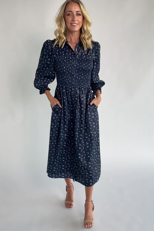 Navy floral button-down midi dress with a collared neckline, smocked bodice, three-quarter sleeves, elastic back waist, side pockets, and a flowing skirt.