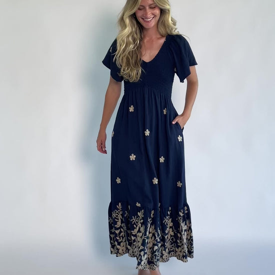 Model wearing a navy embroidered maxi dress with a V-neckline, flutter sleeves, and a smocked bodice.