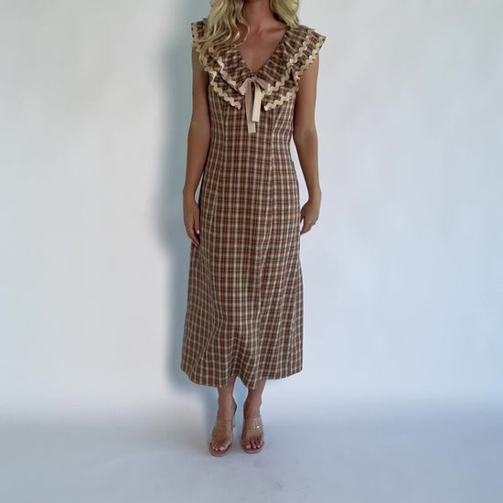 Model wearing a dusty mocha and cream plaid midi dress with a V-neckline, decorative bow tie detail, cascading ruffle trim, and a fitted silhouette.