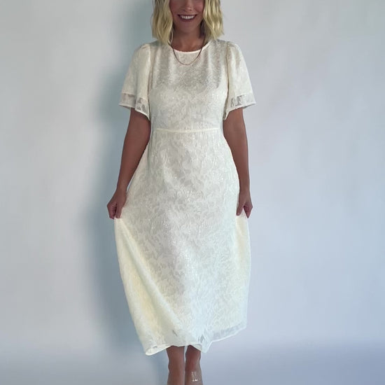 Model wearing a cream midi dress with a round neckline, short gathered sleeves, and a fitted bodice.