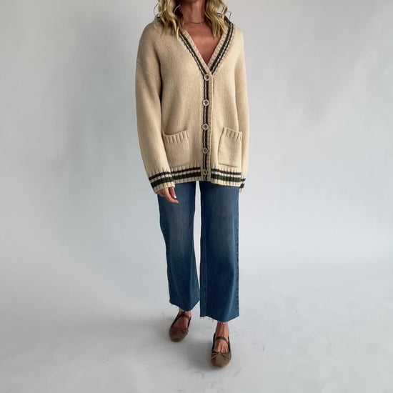 Oatmeal knit cardigan with a V-neckline, button-front closure, olive and charcoal striped trim, front patch pockets, and a relaxed fit.