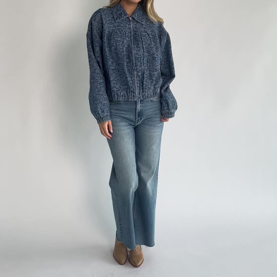 Medium wash paisley print denim jacket with a collared neckline, front zipper closure, long sleeves, elastic hem, and side pockets.
