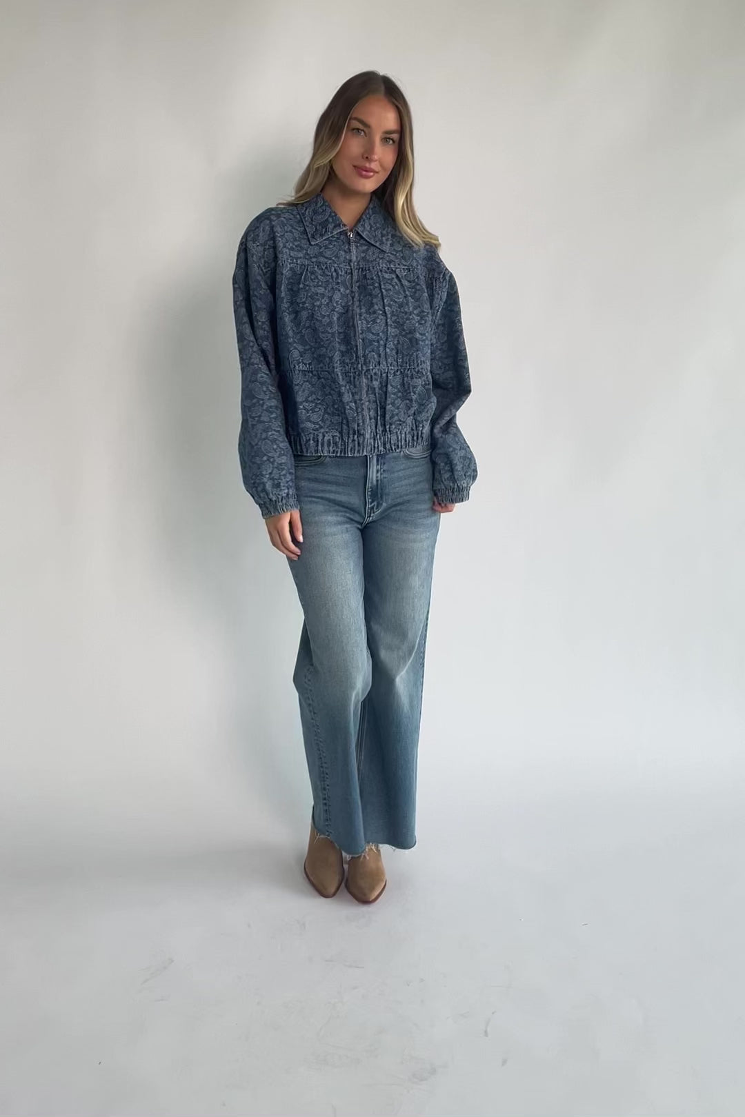 Medium wash paisley print denim jacket with a collared neckline, front zipper closure, long sleeves, elastic hem, and side pockets.