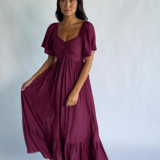 Anisa Midi Dress in Burgundy with a wide V-neckline, cinched bust, elastic shoulders, and a tiered skirt.