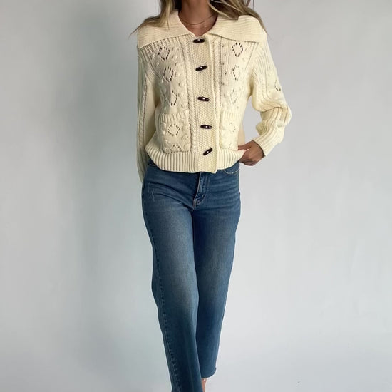 Cream cable knit sweater with a wide fold-over collar, toggle button front, long sleeves, bobble and pointelle knit detailing, ribbed trim, and a relaxed fit.