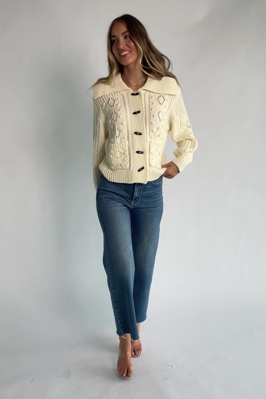 Cream cable knit sweater with a wide fold-over collar, toggle button front, long sleeves, bobble and pointelle knit detailing, ribbed trim, and a relaxed fit.