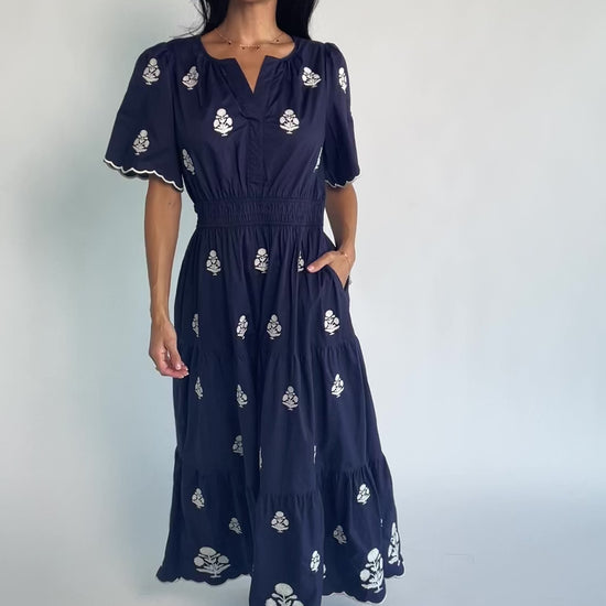 Model wearing the Delilah Embroidered Maxi Dress in navy featuring off-white floral embroidery, a split V-neckline, scalloped short sleeves, a smocked waistband, and a flowing tiered maxi skirt with a scalloped hem.