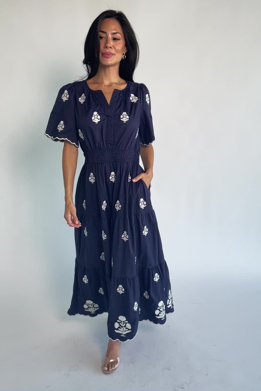 Model wearing the Delilah Embroidered Maxi Dress in navy featuring off-white floral embroidery, a split V-neckline, scalloped short sleeves, a smocked waistband, and a flowing tiered maxi skirt with a scalloped hem.