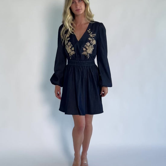 Model wearing a dark chambray short dress with a V-neckline, floral embroidery at the bust, long sleeves with elastic cuffs, and a double elastic waistband.