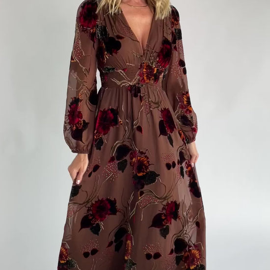 Brown burnout velvet maxi dress with a deep V-neckline, long sleeves, smocked waistband, and flowing asymmetrical hem with gold and burgundy floral accents.