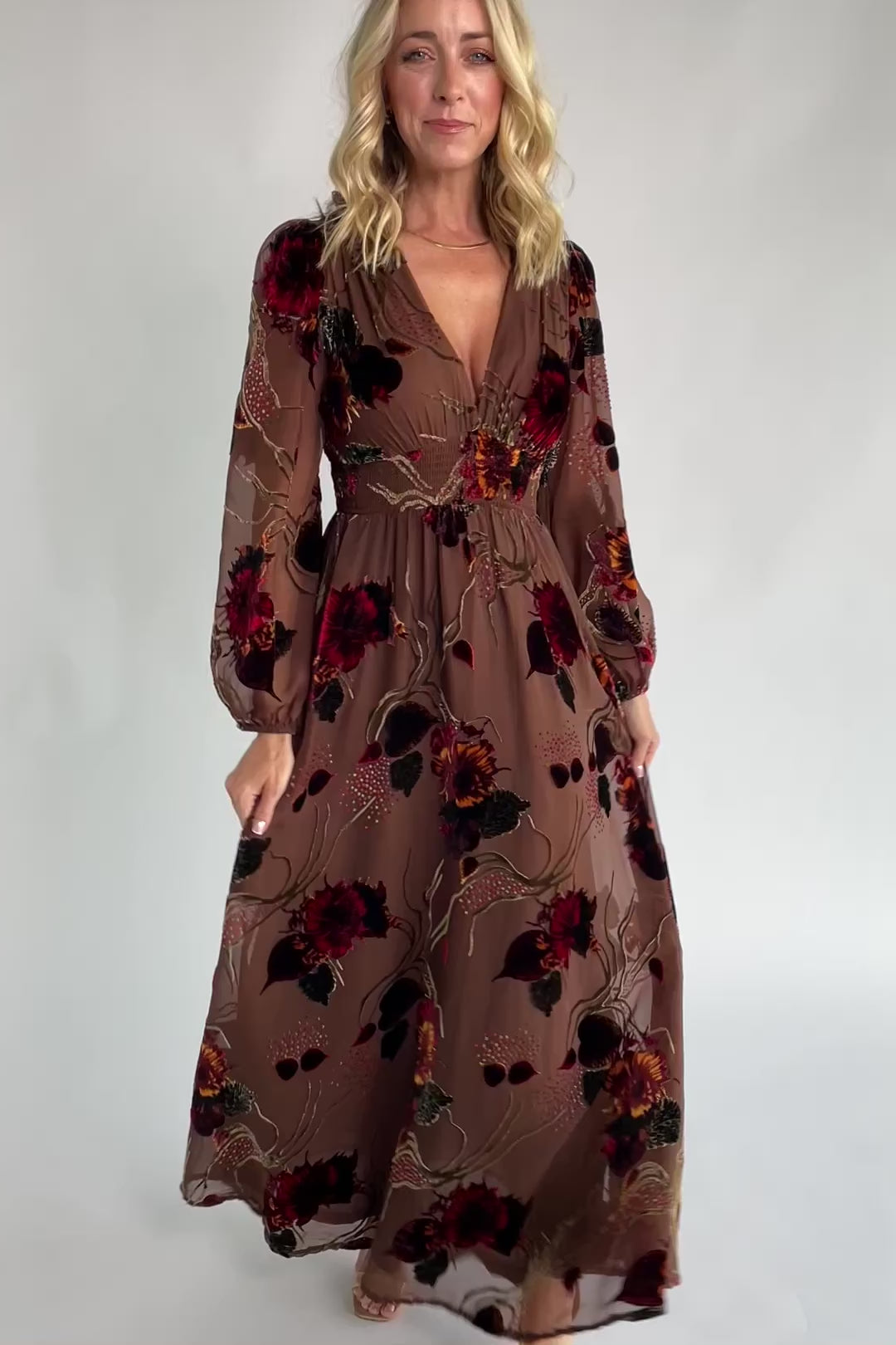 Brown burnout velvet maxi dress with a deep V-neckline, long sleeves, smocked waistband, and flowing asymmetrical hem with gold and burgundy floral accents.