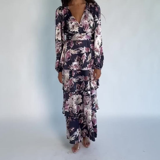 Model wearing the Montrouge Tiered Satin Maxi Dress in Navy + Lilac Floral featuring a V-neckline, short flutter sleeves, a smocked back, and a flowing tiered satin maxi skirt with a navy background and lilac floral print.